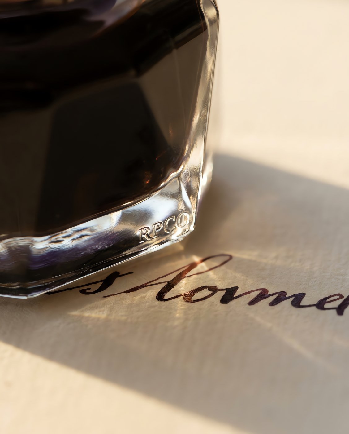 Sepia Copy Ink, 50 ml — A faceted glass bottle of warm brown-black ink on cream paper showing a written line that reads red in the thin strokes in close detail.