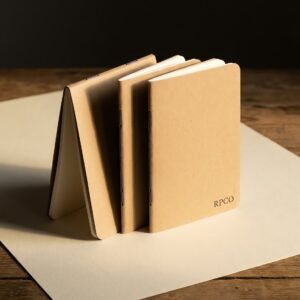 Quire Pocket Notebook, Three Pack — Three slim saddle-stitched pocket notebooks with plain kraft covers fanned out on a wooden desk, one folded fully back on itself.