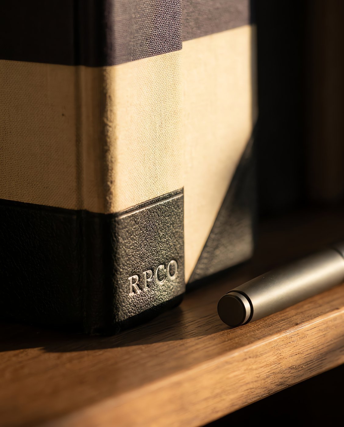 Ledger Book, Half-Bound — A thick half-bound ledger book with a rounded backed spine, cloth boards and leather-grain corners, standing on a shelf beside a closed pen in close detail.