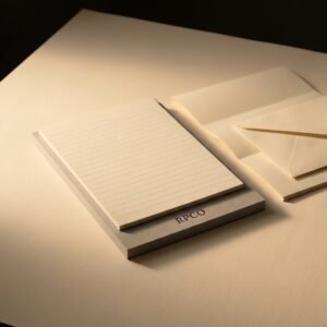Register Pad, A5 — A small A5 writing pad of cream ruled laid paper on a grey board backing, lying beside a folded sheet and an envelope.
