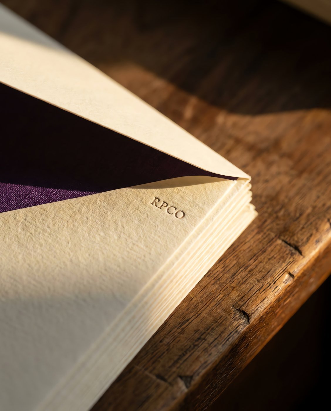 Lined Envelope Box, Fifty — A stack of cream laid envelopes with one flap lifted to show a violet tissue lining, on a wooden desk in close detail.