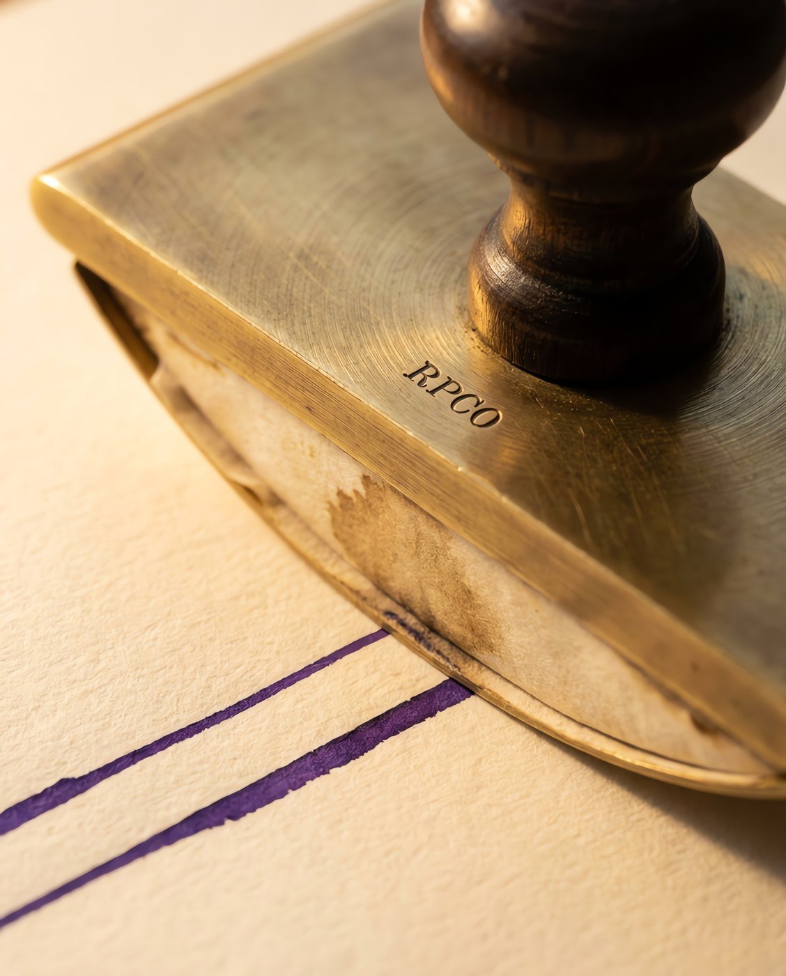 Brass Rocker Blotter — A heavy curved brass rocker blotter with a turned handle, holding a stained cream blotting sheet, rolling across a freshly written page in close detail.