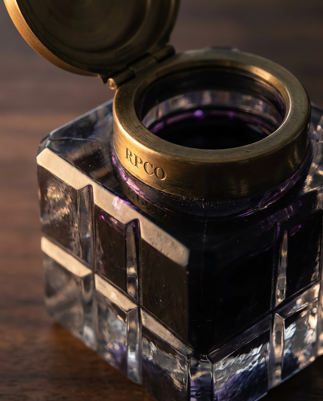 Cut-Glass Inkwell, Brass Collar — A heavy faceted cut glass inkwell with a hinged brass collar and lid standing open on a walnut desk, dark violet ink inside in close detail.