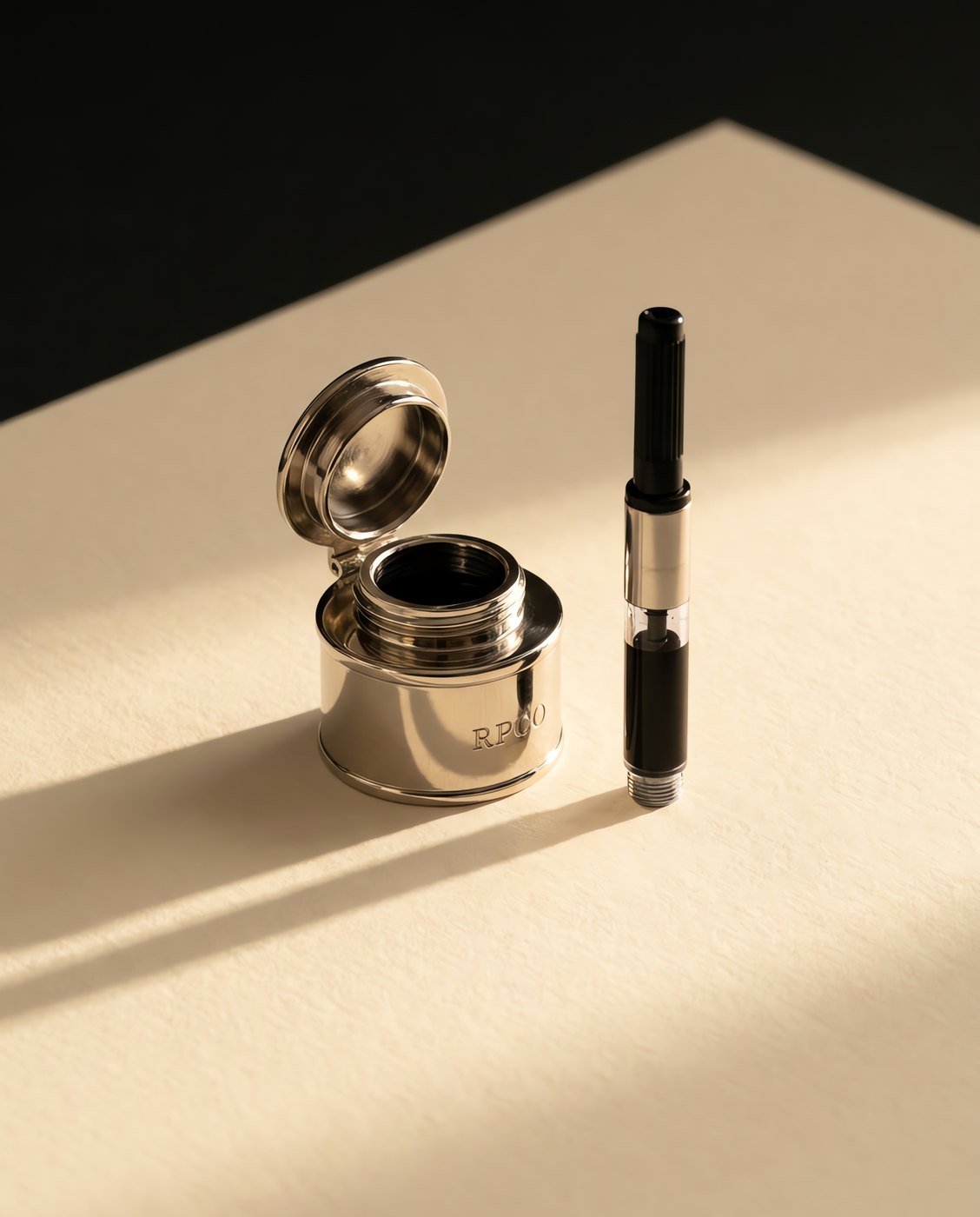 Travelling Inkwell, Nickel — A small cylindrical nickel plated metal inkwell with a bayonet lid, standing open beside a pen converter drawing ink from it.