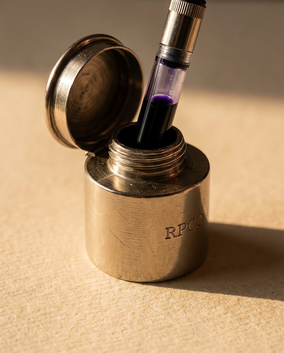 Travelling Inkwell, Nickel — A small cylindrical nickel plated metal inkwell with a bayonet lid, standing open beside a pen converter drawing ink from it in close detail.