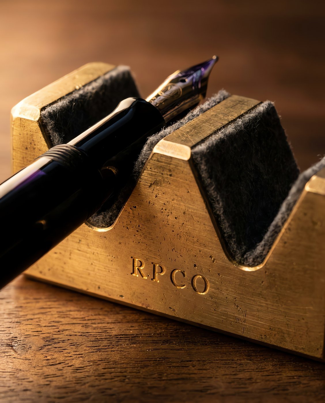 Pen Rest, Machined Brass — A small heavy machined brass pen cradle with two felt-lined vee saddles holding a fountain pen nib-up on a walnut desk in close detail.