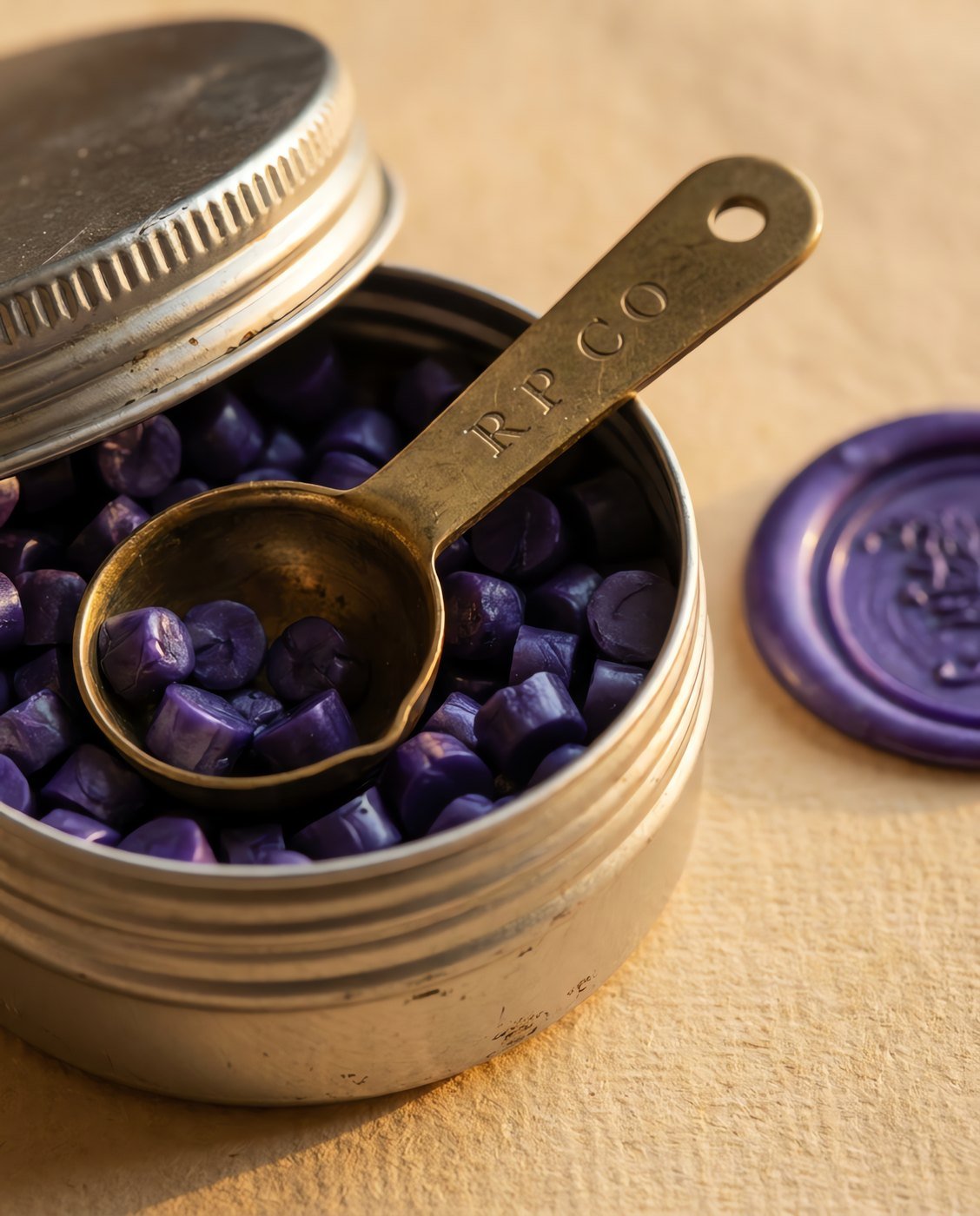 Sealing Wax Beads, Violet, 100 g — An open screw-top tin of small violet wax beads with a small brass measuring scoop, beside a finished wax seal in close detail.