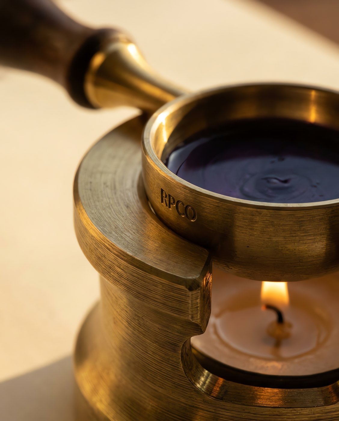 Melting Spoon and Stand — A deep brass melting spoon with a walnut handle resting in a machined brass stand above a lit tealight, violet wax molten in the bowl in close detail.