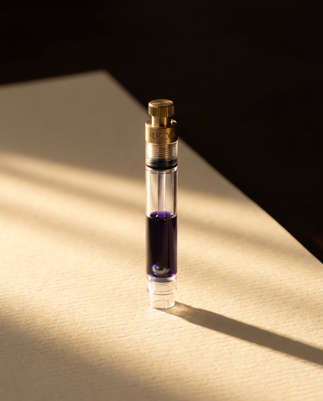 Converter, Piston — A clear standard international piston converter with a brass piston nut, half filled with violet ink, a small steel ball visible inside.