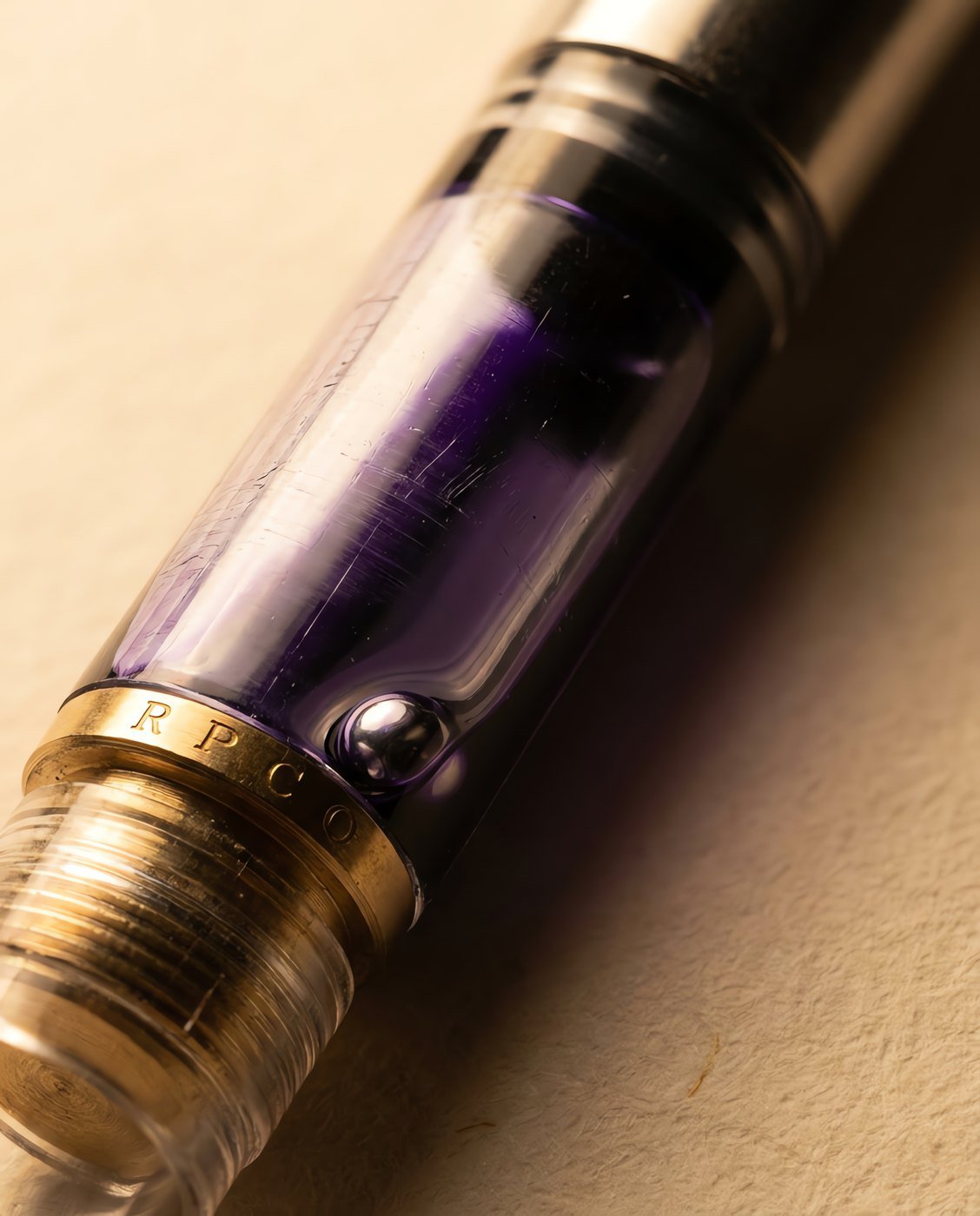 Converter, Piston — A clear standard international piston converter with a brass piston nut, half filled with violet ink, a small steel ball visible inside in close detail.