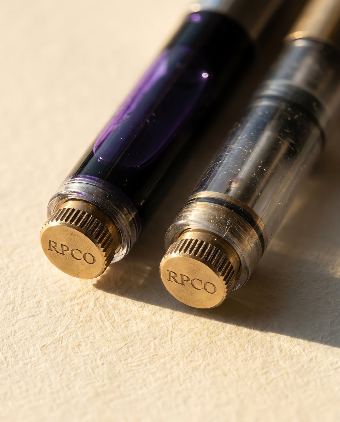 Converter, Twin Pack — Two clear piston converters with brass nuts lying side by side on a card sleeve, one filled with violet ink and one empty in close detail.